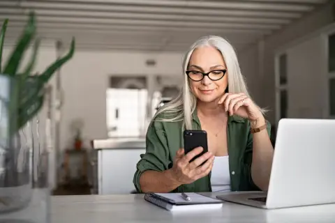 Frau mit Brille und Handy am Tisch, plant das Haus verkaufen und in Mietwohnung ziehen.
