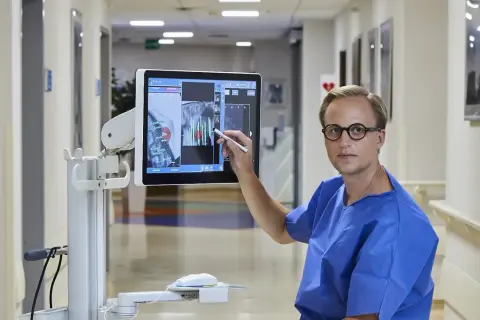 Lekarz w niebieskiej koszuli wskazuje na ekranie komputerowym obraz z planowaniem biopsji gruczołu krokowego.