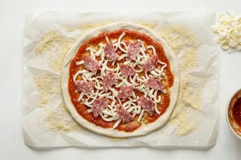 Okrągłe ciasto na pizzę z sosem, serem i salami, przygotowane na papierze do pieczenia.