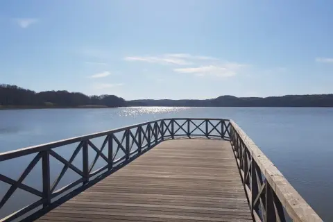 Holzsteg am Schmachter See lädt zum Spaziergang auf dem Rundweg ein. Sonnenschein glitzert auf dem Wasser, bewaldete Ufer im Hintergrund.