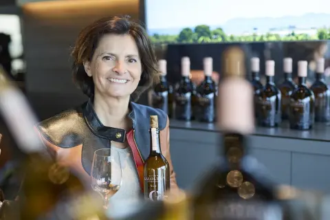 Frau mit Weinflasche und Glas, im Hintergrund Flaschenreihe und Landschaft.