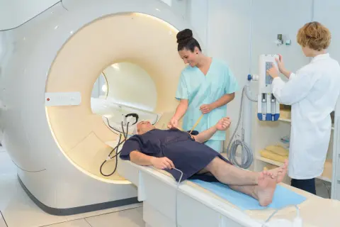 Pacjent leży na łóżku przed aparatem MRI. Dwie pielęgniarki przygotowują go do badania. To właśnie mri co to za badanie.