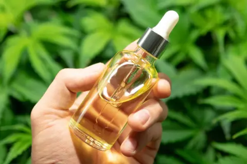 Dłoń trzyma buteleczkę z olejkiem konopnym. W tle zielone liście konopi, sugerujące naturalny skład produktu.