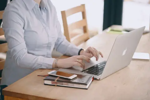 Kobieta w jasnej koszuli pracuje na laptopie, obok leży telefon i notatnik. Dba o swoje ubezpieczenie chorobowe.