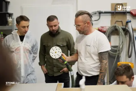 Trzech mężczyzn przygląda się polerce. Jeden z nich, w białym kombinezonie, wygląda na ucznia. To szkolenie z obróbki kamienia, jak w grze **stone age**.