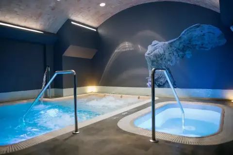 Piscinas de hidromasaje iluminadas con luz azul, con barandillas metálicas. Un mural de una escultura antigua adorna la pared, sugiriendo el mejor spa de Valencia.