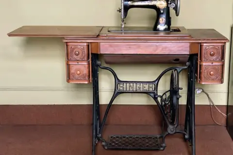 Antique Singer sewing machine on a wooden table with drawers and a treadle base.