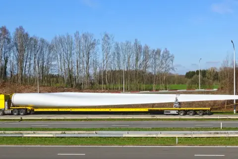 Żółta ciężarówka transportuje ogromne łopaty wiatraka. Ten pojazd nienormatywny pokonuje drogę, by dostarczyć element do budowy farmy wiatrowej.