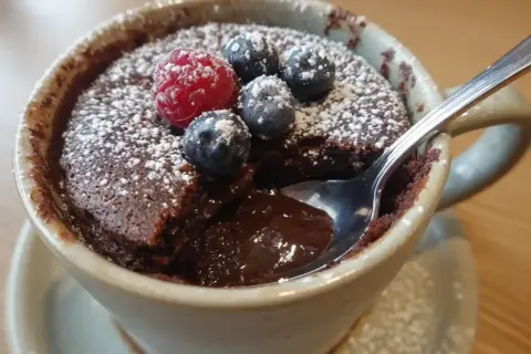 Chokladig mug cake med bär och florsocker. En sked dyker ner i den smältande chokladen.