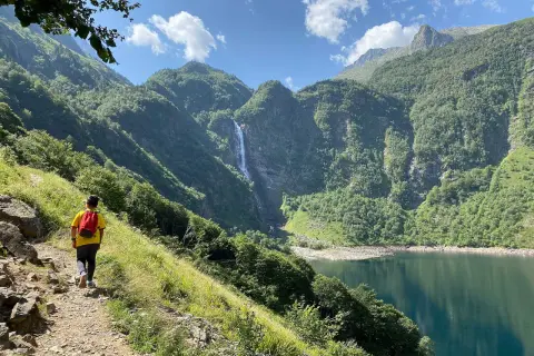Randonneur admirant la cascade et le lac de Toulouse, entourés de montagnes verdoyantes sous un ciel bleu.