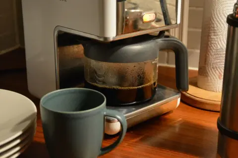 En kaffebryggare med en glaskanna full med färskt kaffe. Bredvid står en mugg och en stapel tallrikar. Man kan brygga presskaffe i kaffebryggare för en god start på dagen.