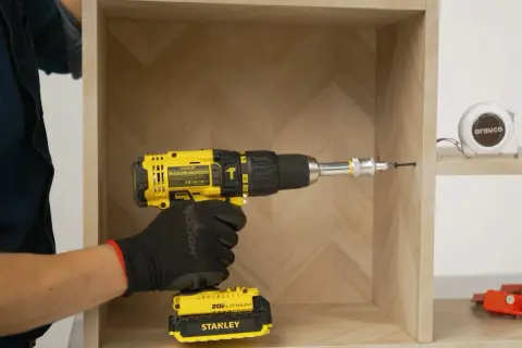 Mano con guante negro usa taladro amarillo para unir dos muebles. Cinta métrica y herramientas al fondo.