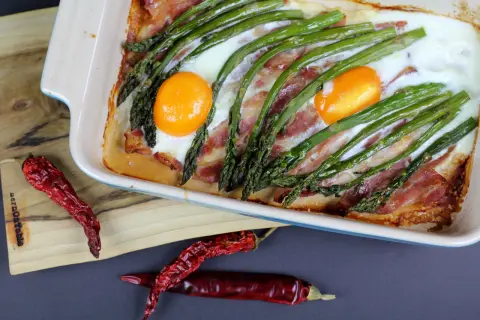 Pyszne śniadanie ze szparagami, jajkami i boczkiem zapieczone w naczyniu. Obok suszone papryczki chili.