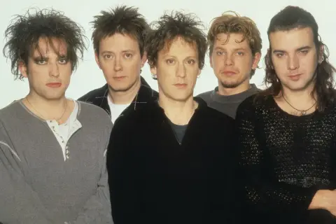 Pięciu mężczyzn, członkowie zespołu The Cure, pozuje do zdjęcia. Ich charakterystyczne fryzury i makijaż podkreślają mroczny klimat ich muzyki.