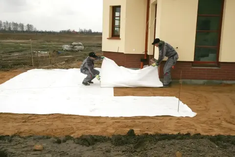 Dwóch mężczyzn rozkłada białą geowłókninę, przygotowując tani taras.