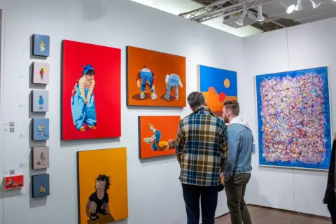 Two men examine colorful paintings at an art fair, perhaps contemplating their next acquisition as an art collector.