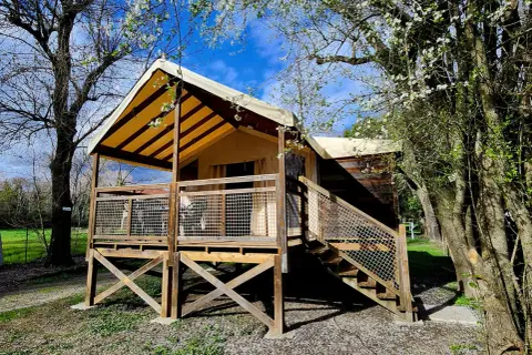 Un lodge de camping éco-responsable avec une tente surélevée et une terrasse en bois, entouré d'arbres fleuris sous un ciel bleu.