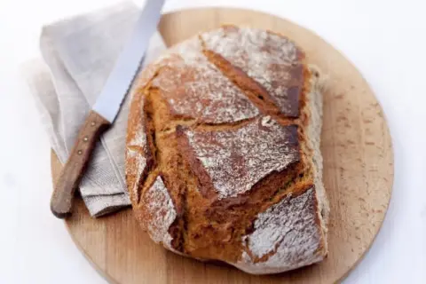 Ein frisch gebackenes Brot aus Hefe Teig liegt auf einem Holzbrett. Daneben ein Messer und eine Serviette.