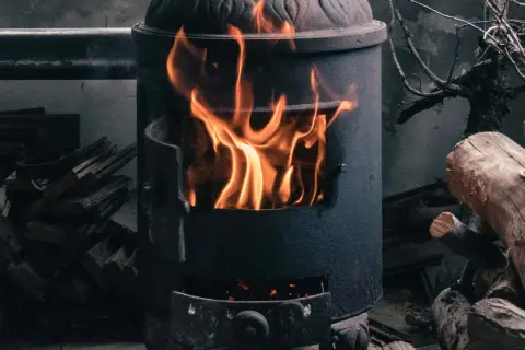 Ein Kaminofen brennt hell. Achte auf ausreichende Belüftung, um Sauerstoffmangel durch Kaminofen zu vermeiden. Holzscheite liegen daneben.