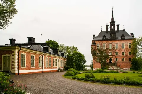 Vackert vik slott med en lång, röd tegelbyggnad och ett torn med klocka. Grönskande trädgård och en grusväg leder fram till byggnaderna.
