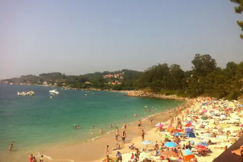 Gente disfrutando del sol y el mar en la playa areacova, con barcos en el agua y colinas verdes al fondo.