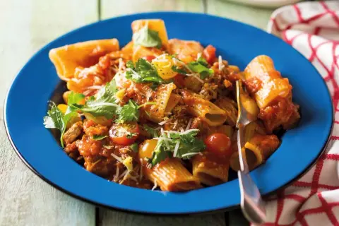 Pyszne mezzi rigatoni w sosie pomidorowym z mięsem mielonym, pomidorkami koktajlowymi i świeżą natką pietruszki, podane na niebieskim talerzu.
