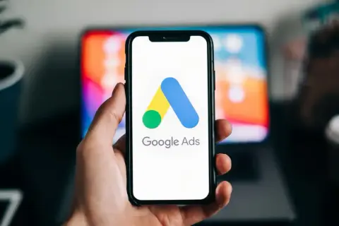 Dłoń trzyma smartfon z logo Google Ads. W tle widać ekran laptopa. Dziś budżet w reklamie Google Ads ustalamy.