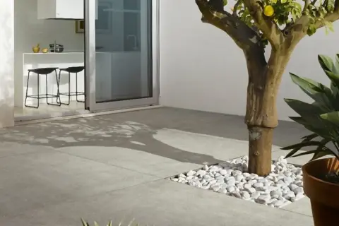 Un moderno patio con diversi tipi di pavimentazione esterna: lastre in gres grigio e un'area con ciottoli bianchi attorno a un albero.