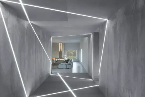 Nowoczesny korytarz z betonowymi ścianami i sufitem, ozdobiony geometrycznymi liniami światła LED. W tle widać salon z kominkiem i meblami. To inspiracja, jak zamontować oświetlenie LED w witrynie.