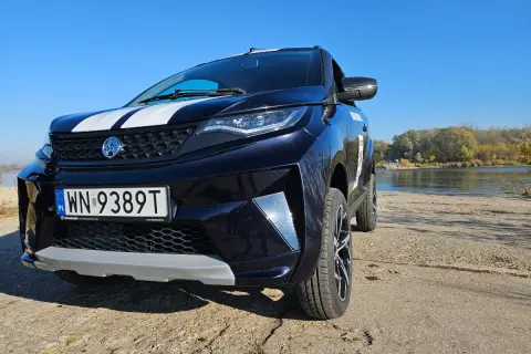 Ciemnoniebieski SUV z białymi pasami na masce, zaparkowany nad rzeką. To auto od 14 lat, gotowe na przygodę.