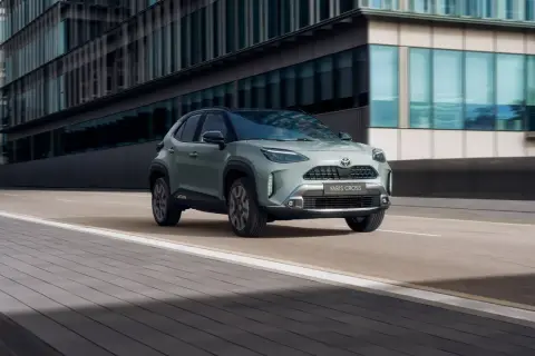 En grön Toyota Yaris Cross hybrid kör på en stadsgata. Bilen är i fokus framför moderna byggnader.
