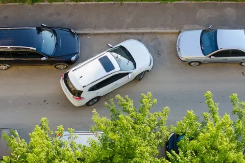 Biały SUV wykonuje parkowanie równoległe przodem między czarnym SUV-em a srebrnym sedanem.