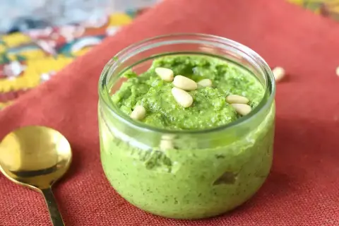 En burk med hemgjord egen pesto, toppad med pinjenötter. En guldig sked ligger bredvid på en röd duk.