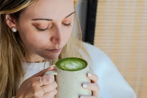 Femme aux cheveux blonds savourant un matcha latte crémeux, le regard clos. La boisson verte présente un joli dessin de mousse.