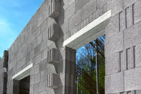 Ściana z bloczków betonowych z widocznym betonowym nadprożem nad otworem okiennym. W tle zbrojenie i drzewa.