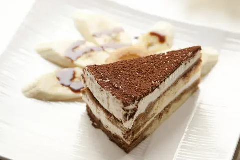 Tranche de tiramisu banane, saupoudrée de cacao, accompagnée de rondelles de banane nappées de sauce. Un délice !