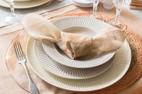 Une table dressée avec des assiettes empilées, une serviette en tissu nouée, une fourchette et des verres. La différence entre faïence et porcelaine est subtile, mais la qualité est évidente.