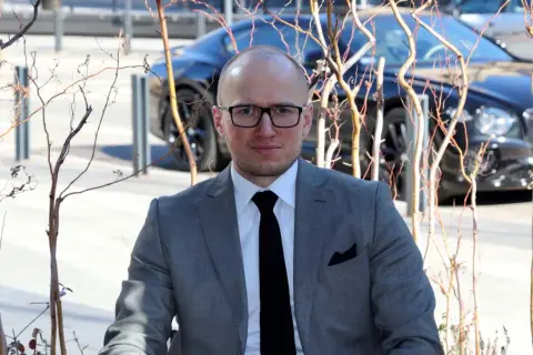 Mężczyzna w garniturze, z okularami, na tle luksusowego samochodu. Jego styl sugeruje sukces i przemyślane inwestycje rzeczowe.