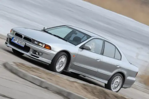 Srebrny Mitsubishi Galant z kierowcą za kierownicą, pokonujący zakręt na drodze.