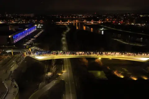Nocne życie tętni na mostach berdychowskich. Tłumy ludzi spacerują po oświetlonym moście, a w tle widać miasto i rzekę.