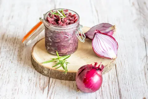Słoik z konfiturą z czerwonej cebuli, obok cała cebula i połówka. Gałązka rozmarynu dodaje aromatu.