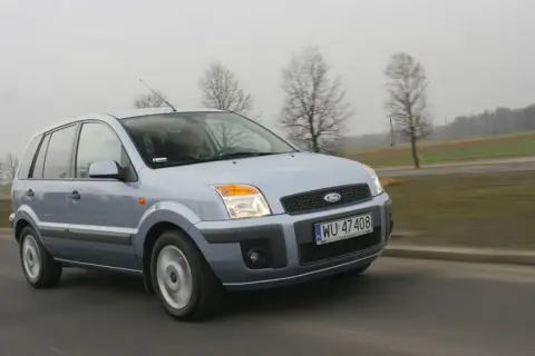 Jasnoszary Ford Fusion 1.4 benzyna jedzie po drodze. W tle drzewa i pola.