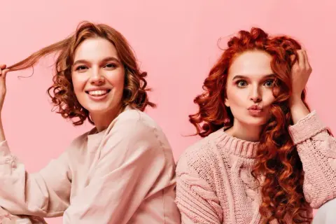 Dos chicas con diferentes tipos de cabello ondulado, una sonríe y juega con su pelo, la otra hace un puchero.