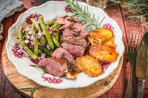 Saftiger Lammbraten nach Omas Rezept, serviert mit grünen Bohnen, roten Zwiebeln und knusprigen Kartoffelscheiben auf einem Teller mit Blumenmuster.