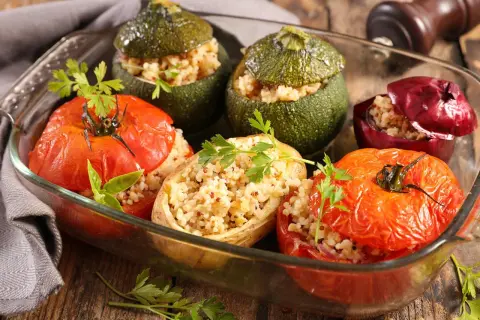 Un plat de petits farcis gastronomiques : tomates, courgettes rondes et oignons rouges farcis de grains et herbes, dans un plat en verre.