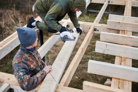 Dwóch mężczyzn buduje konstrukcję z drewna. Jeden z nich używa piły tarczowej do cięcia belek, które są częścią projektu.