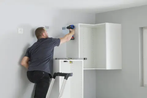 Hombre en escalera usa taladro para fijar soportes en pared, parte de como colocar kit para colgar armarios.