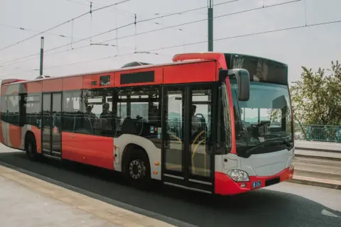 Czerwony autobus miejski jedzie po moście, w tle widać drzewa i błękitną poręcz. To idealny środek transportu, by dojechać nad morze.