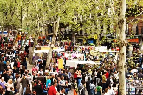 Gente comprando en mercadillos de la sierra de Madrid, con puestos de ropa y accesorios bajo árboles frondosos.