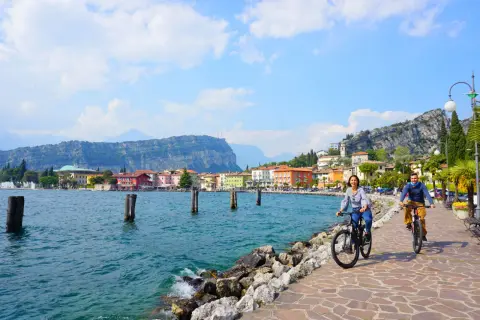 Coppia in bici sul lungolago di Riva del Garda, tra le attrazioni del posto, con case colorate e montagne sullo sfondo.